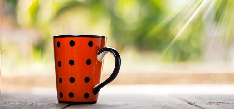 Eine orangefarbene Kaffeetasse mit schwarzen Punkten steht auf einem Holztisch und wird von Sonnenstrahlen beschienen.