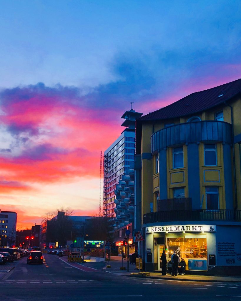 Rechts ein Eckhaus mit einem hell beleuchteten Kiosk mit dem Namen Kesselmarkt. Daneben eine Straße mit Autos und im Hintergrund geht die Sonne unter und färbt die Wolken in tollen Rottönen